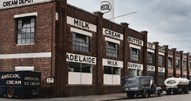 Colourised historic photograph of the Amscol Adelaide Milk Supply Co-op factory, with vintage ice cream and milk delivery trucks parked outside.